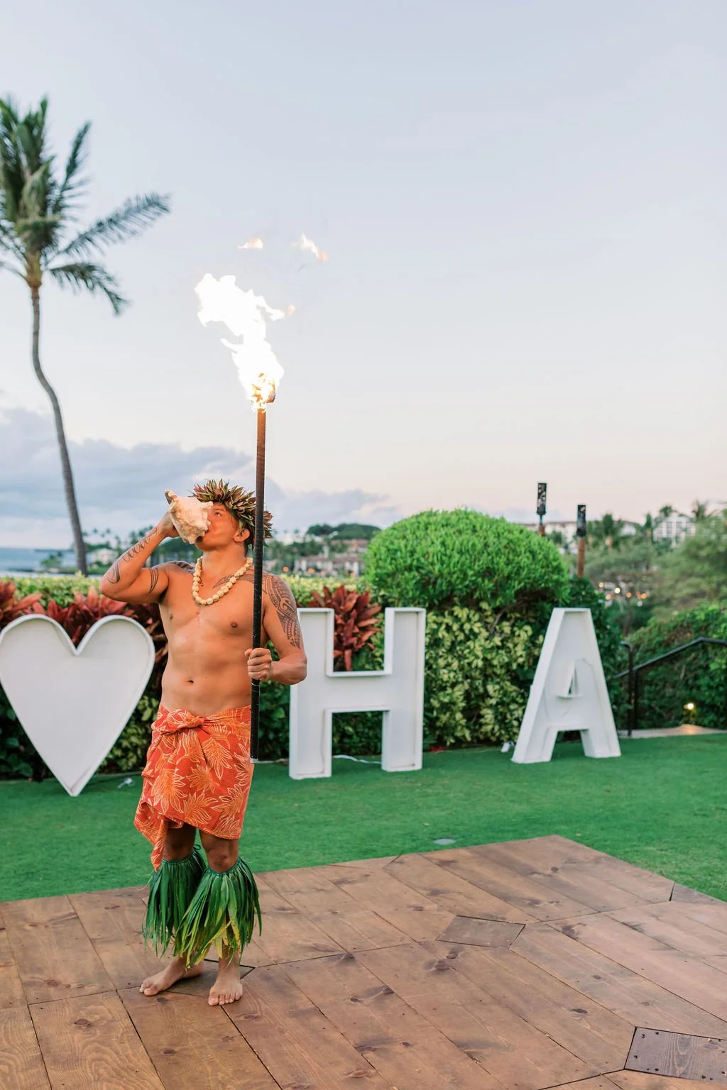 Hawaiian performer with conch shell and torch beside the ALOHA letters