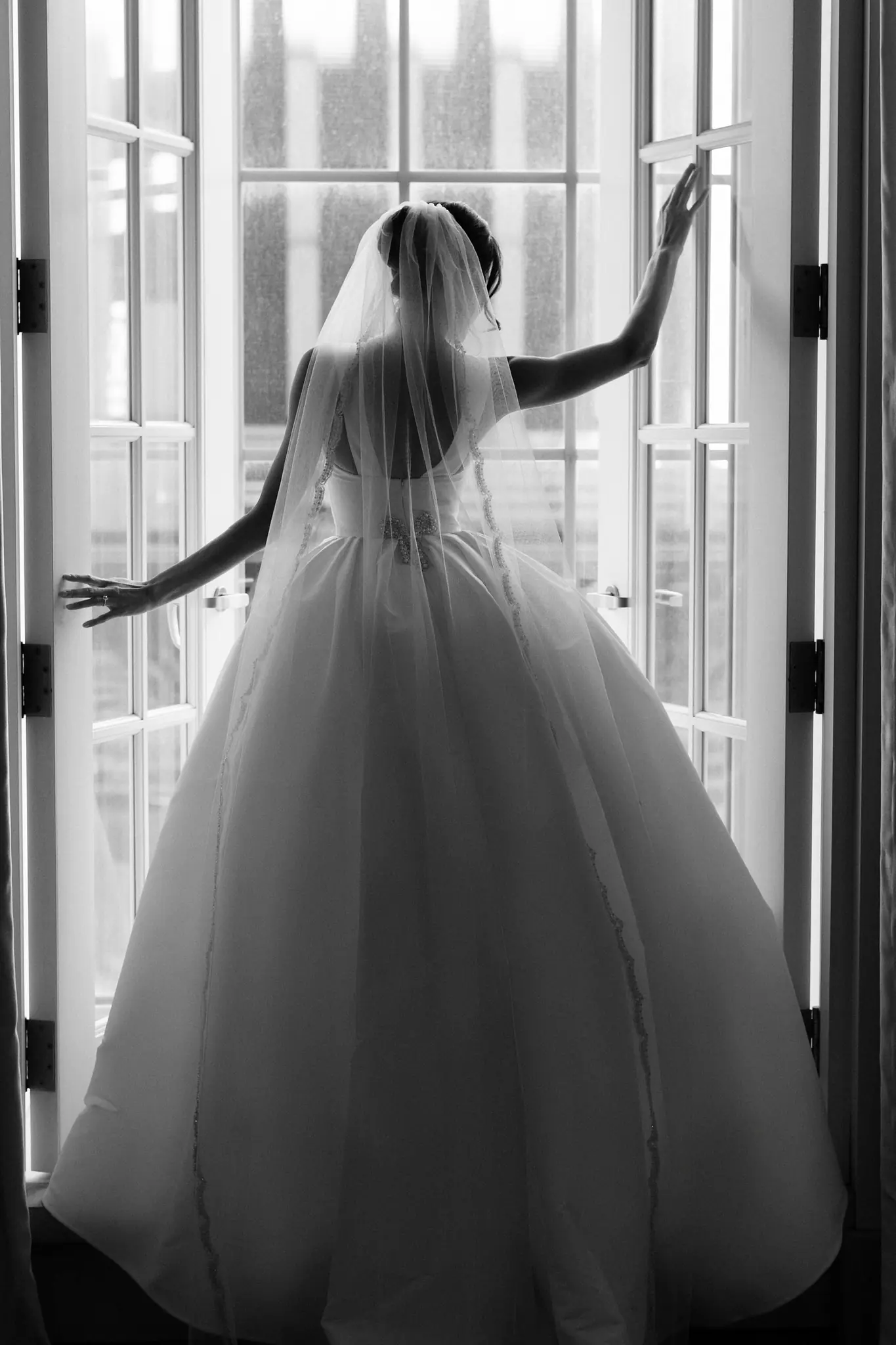 A bride in a cathedral veil silhouetted at open French doors, photographed in black and white