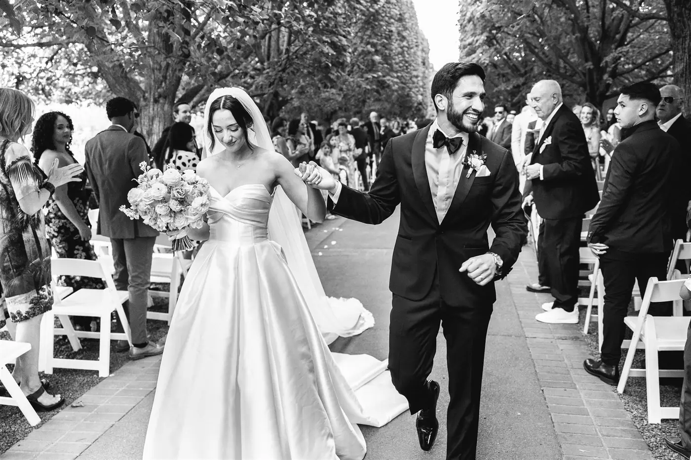 A newly married couple walking the aisle hand in hand beneath a canopy of trees, in black and white