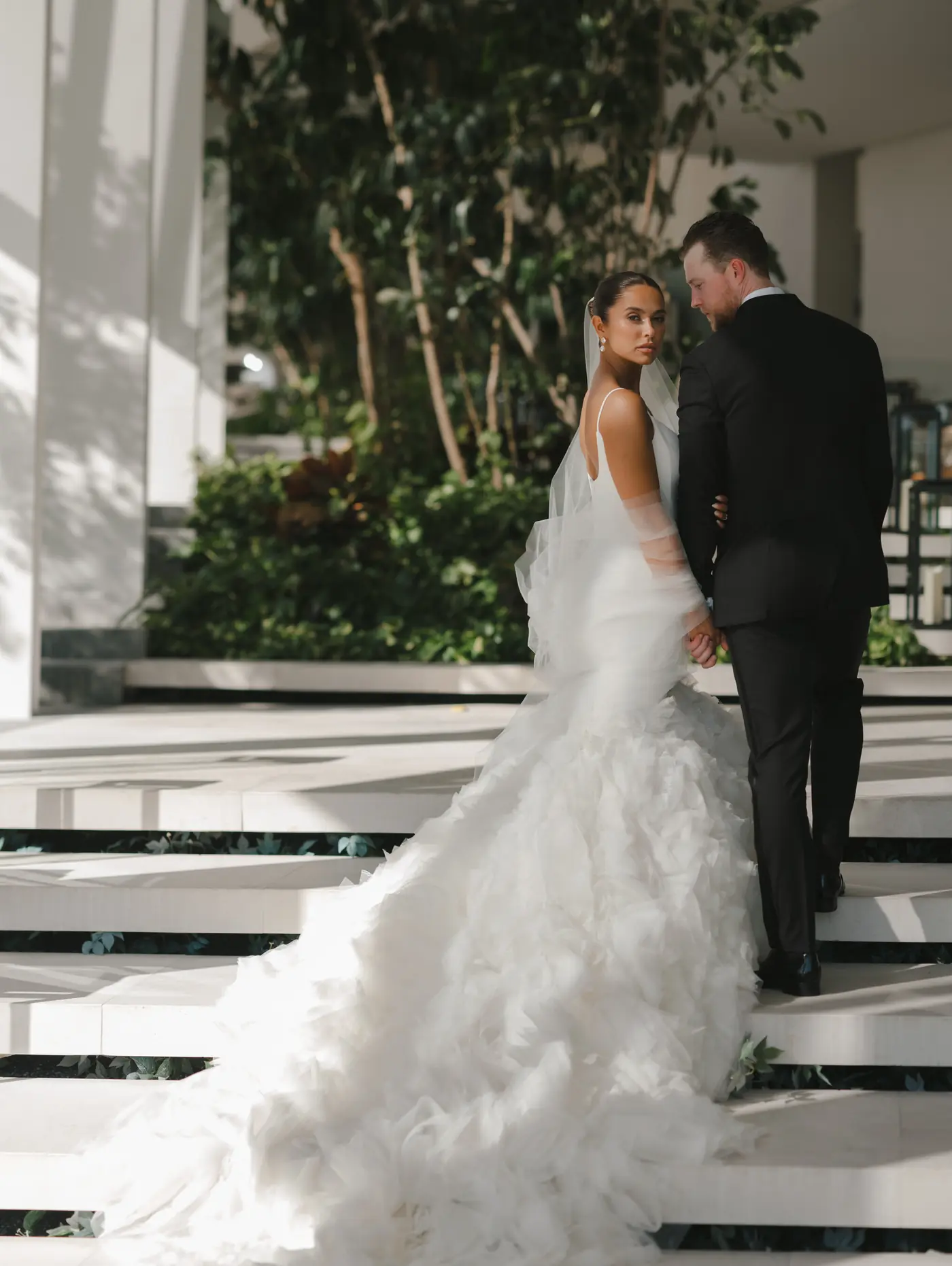 Bride in a feathered gown and veil glancing back while climbing sunlit stone steps hand in hand with the groom at a destination wedding planned by Michigan Avenue Events