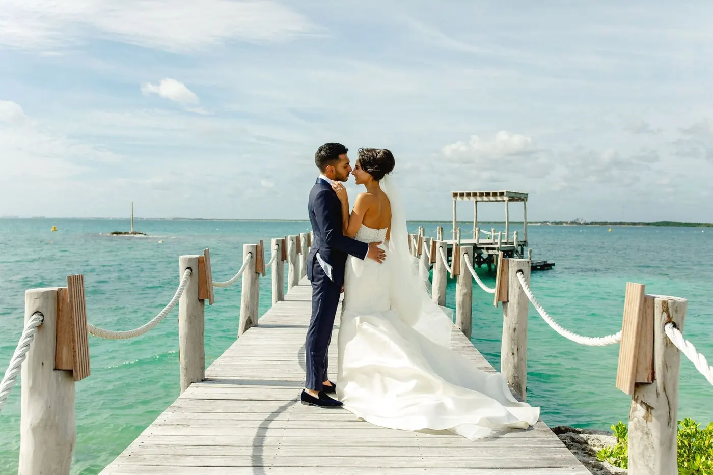 Lara Sofia and Atul embracing on a pier over turquoise water at NIZUC